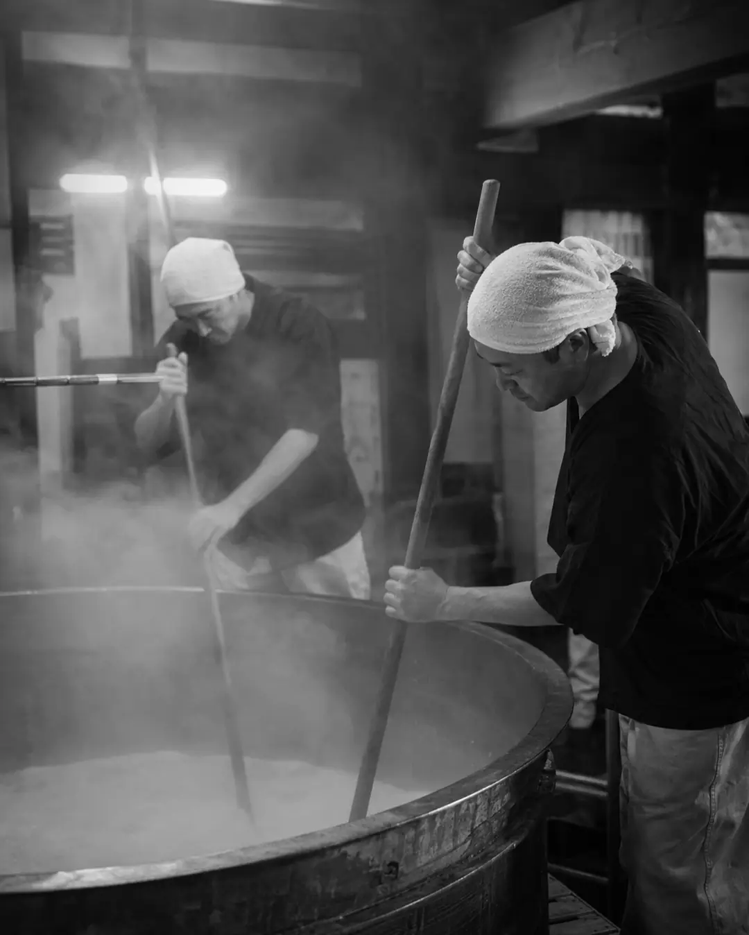 Two brewers stirring a steaming tank of sake mash with long wooden poles