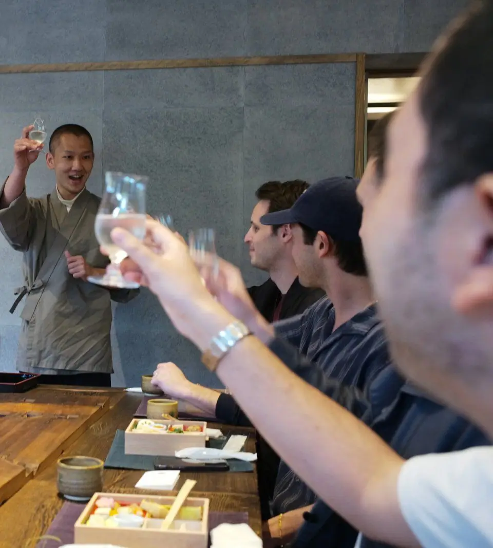 A host raising a glass of sake at a wooden counter as guests toast back